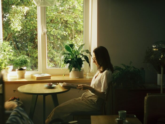A woman reading at a table surrounded by plants, deep in thought as her inner critic lingers in the background.