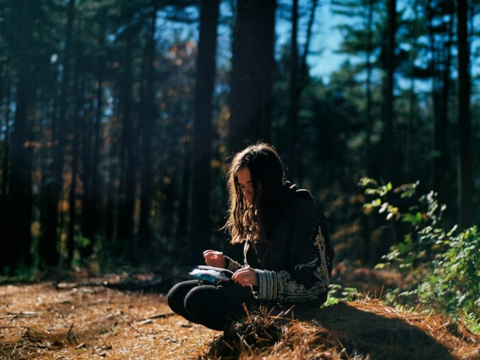 A woman sitting in the woods, writing in a notebook, embodying the tension between inspiration and creative anxiety.